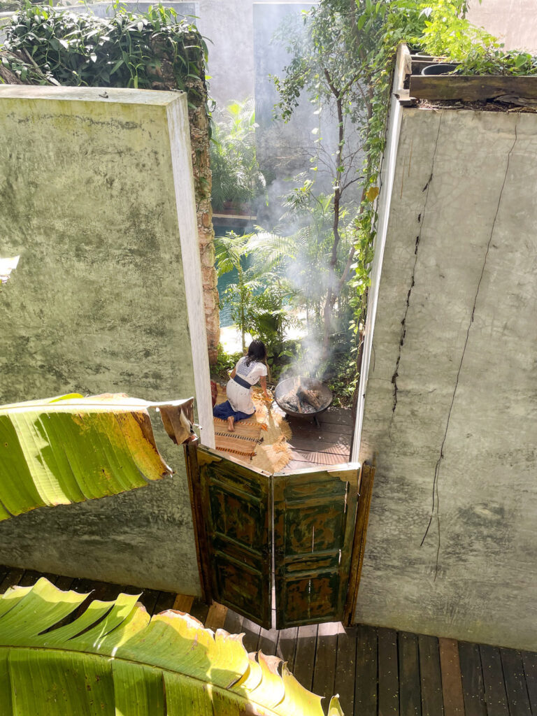 A woman in a white dress kneeling down at a basin stoking a fire.