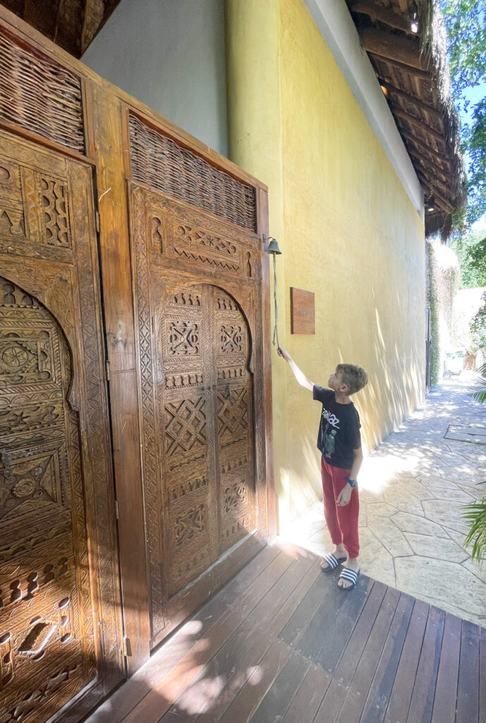 A child ringing the doorbell hanging next to the large, wood carved doors in front of Casa Hormiga Bacalar