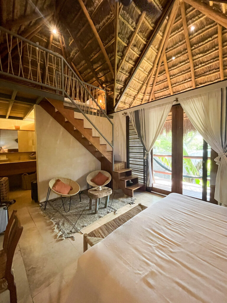 Two chairs, a table, and the foot of the bed visible inside the Tree Top Loft Suite at Casa Hormiga Bacalar.
