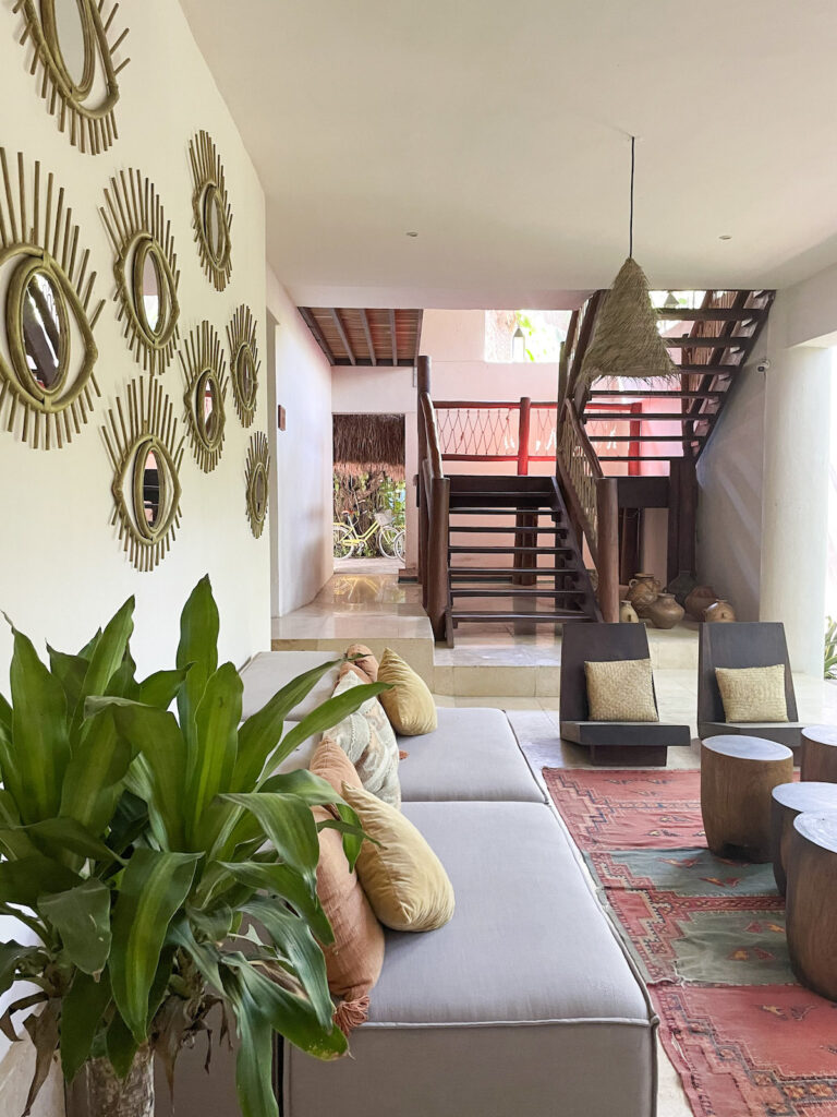 The sitting space featuring a couch, wooden tables and two wooden chairs with cushions backed by a mirrored wall and staircase at Casa Hormiga Bacalar.