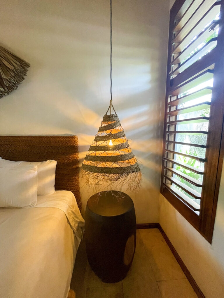 A photo of the edge of a bed, a wooden table and hanging light fixture next to a window.