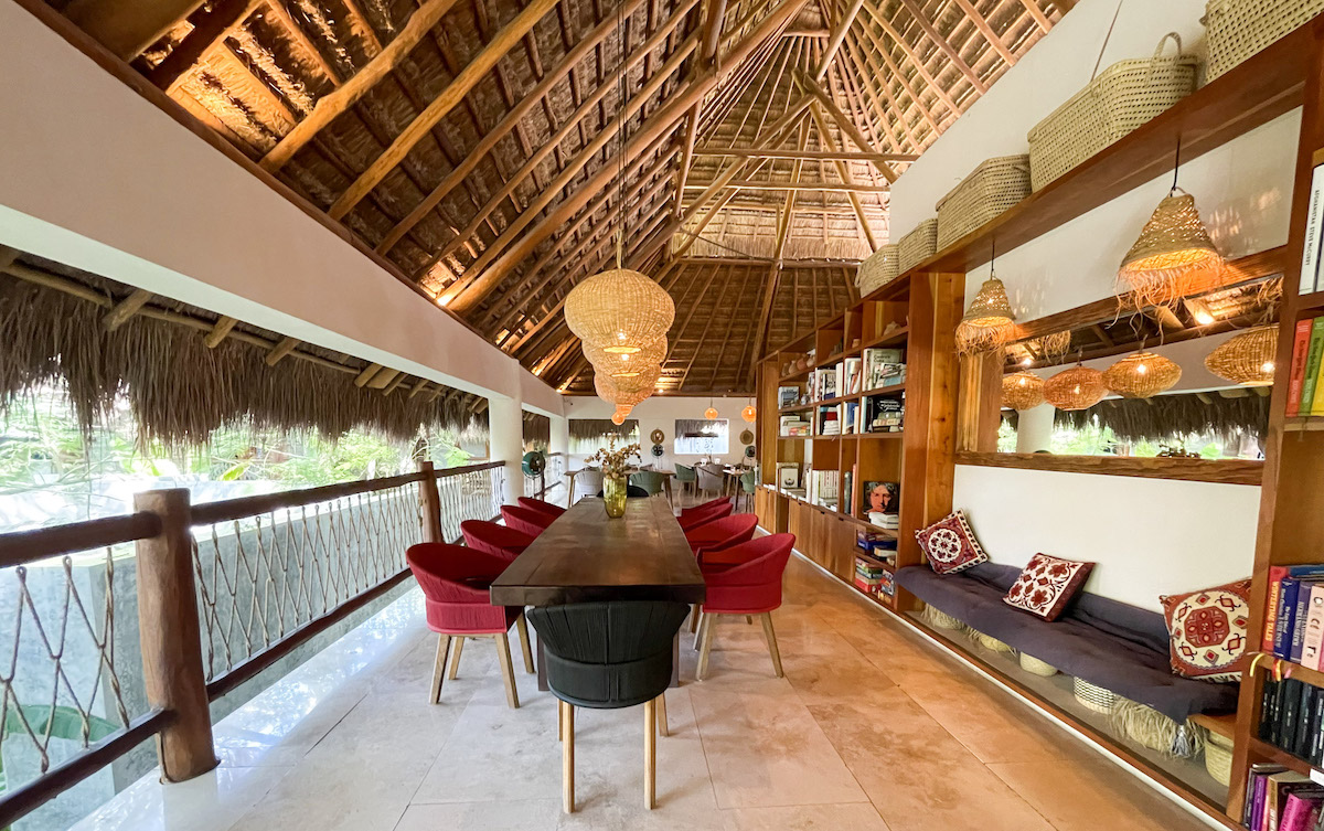 A long table surrounded by burgundy and black chairs that's flanked by a bookcase and bench seating under a palapa roof at Casa Hormiga Bacalar.