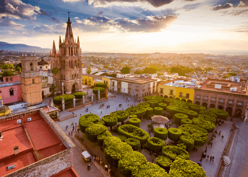An aerial shot of large, Baroque church, a neighboring bell tower and colorful buildings with windows and archways surrounding a verdant city square planted with trees in San Miguel de Allende, a great place to spend summer in Mexico.