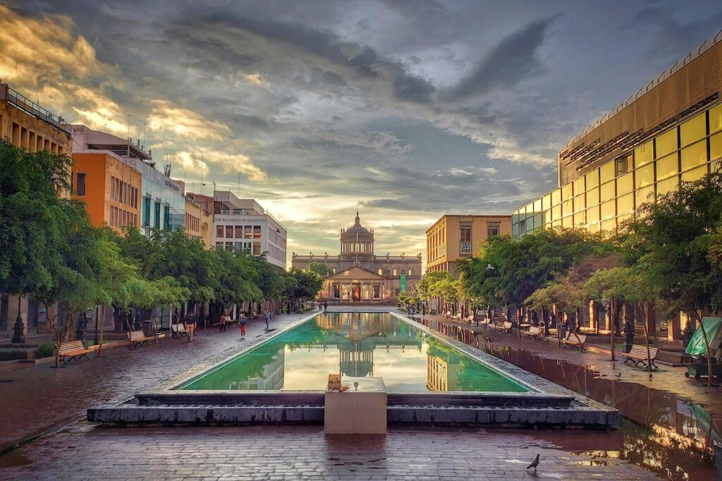 In the center, a long pool of water flanked by modern buildings, some colorful, and some with glass windows, all leading to a neoclassical looking building with a large dome in the distance beneath a cloudy sky in Guadalajara, Mexico.