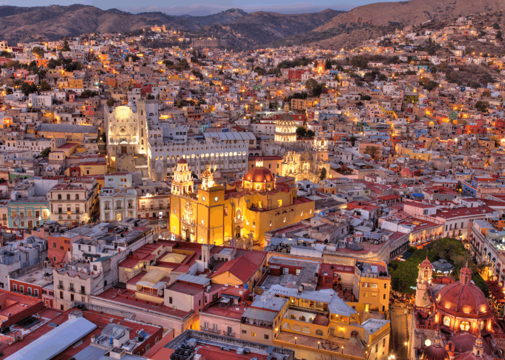 An aerial view of yellow, white and terracotta colored buldings with glowy lights planted over the hills of Guanajuato, Mexico, with mountains visible in the background.