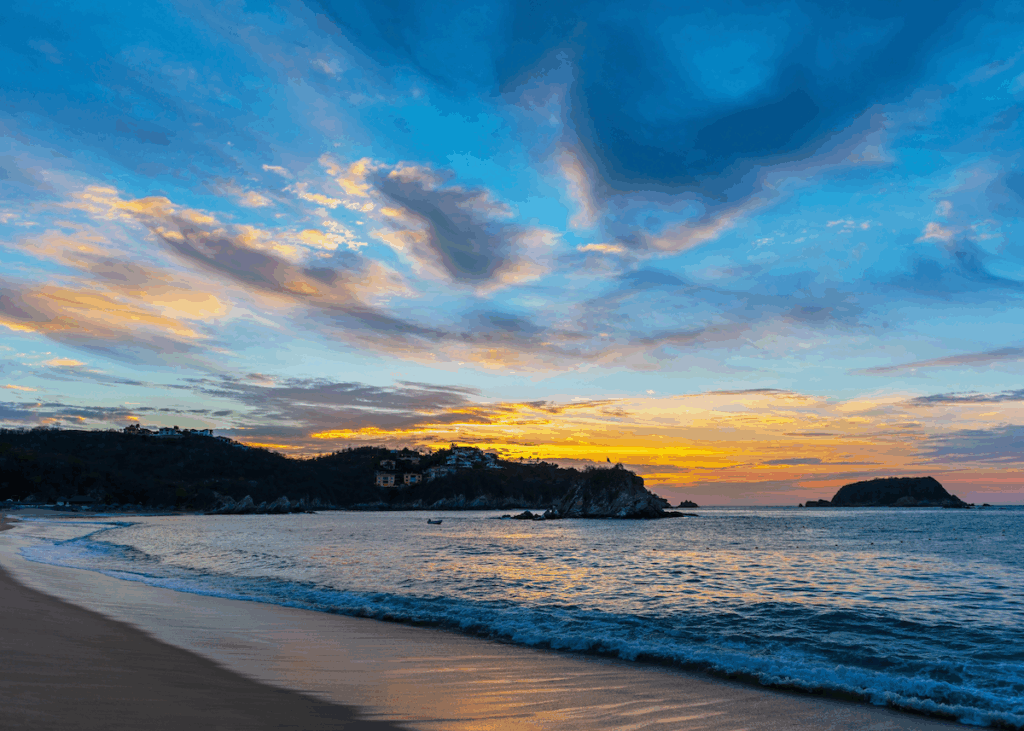 A blazing orange sunset in a cloudy cobalt sky over a lush hillside cradling a gentle ocean rolling onto the sandy shores of Huatulco, Mexico.