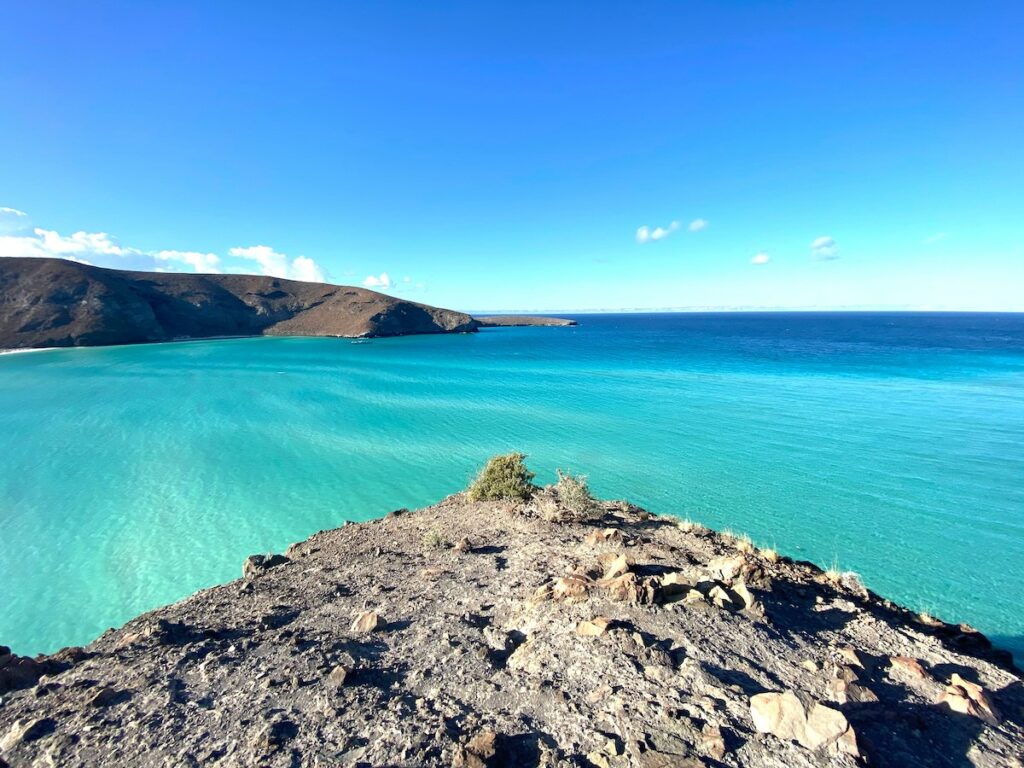 A blazing blue ocean and clear blue sky surrounded by rolling hills in the background and a rocky cliff in the foreground in La Paz, an excellent place to summer in Mexico.
