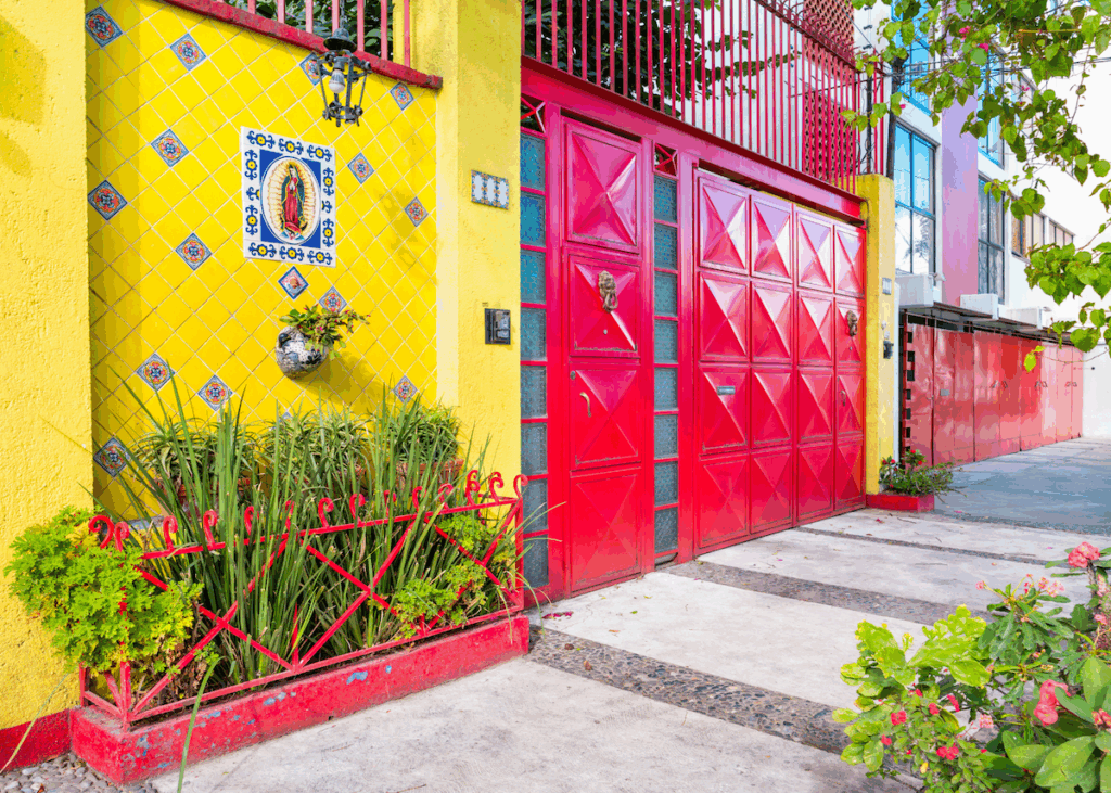 The vibrant yellow and red exterior of a building with tiled walls and metal doors flanked by plants and trees along a street in Mexico City.