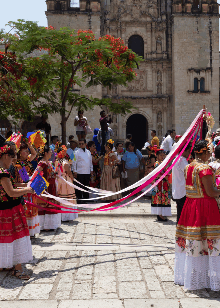 Several women in colorful, traditional Mexican dresses holding red and white streamers as the stand in front of a church in Oaxaca, Mexico.