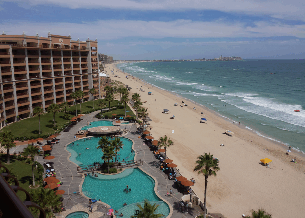 To the left, a large hotel resort with grassy gardens and a pool flanked by terracotta umbrellas in front. The the right, a sandy stretch of beach with the foamy ocean rolling up to shore.