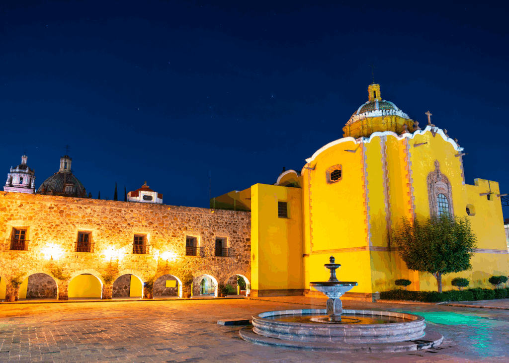 To the right, a long, low wall with domes visible above and behind it, and to the right an ornate, yellow church with an egg shaped dome, all beneath the sky at dusk. In the foreground, a fountain.
